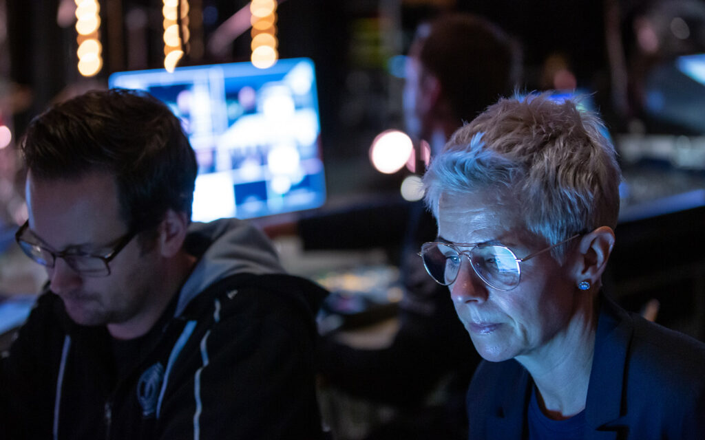 Tekniker tillsammans med Ann Rebenstam bakom kulisserna vid ett event. Monitorer lyser upp i bakgrunden också.