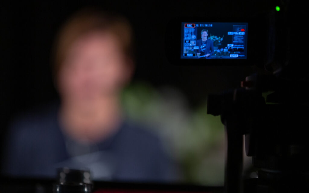 Suddig bild av person på scenen, men på en liten skärm ser man en klar bild av personen på scenen.