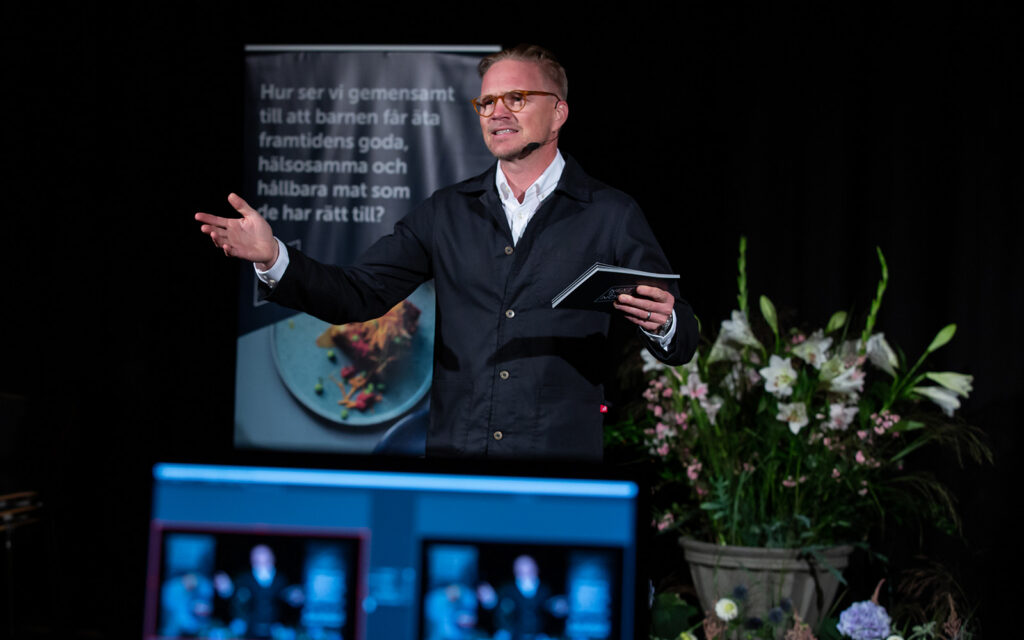 Jens Dolk står med utsträckta armar på scenen och föreläser.