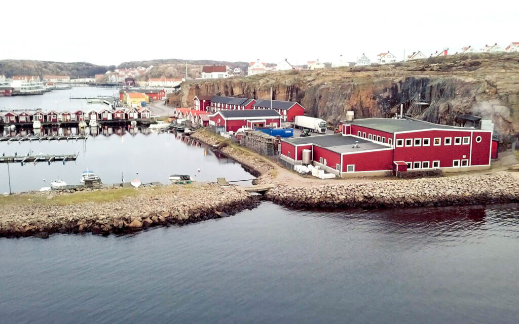 Vy utanför bergrummet. Hav, småbåtshamn, stugor och lite större röda byggnader i anslutning till bergrummet.