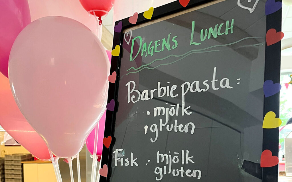 Menyskylt i skolmatsal där det står bland annat Barbiepasta. Rosa ballonger syns vid sidan om skylten.
