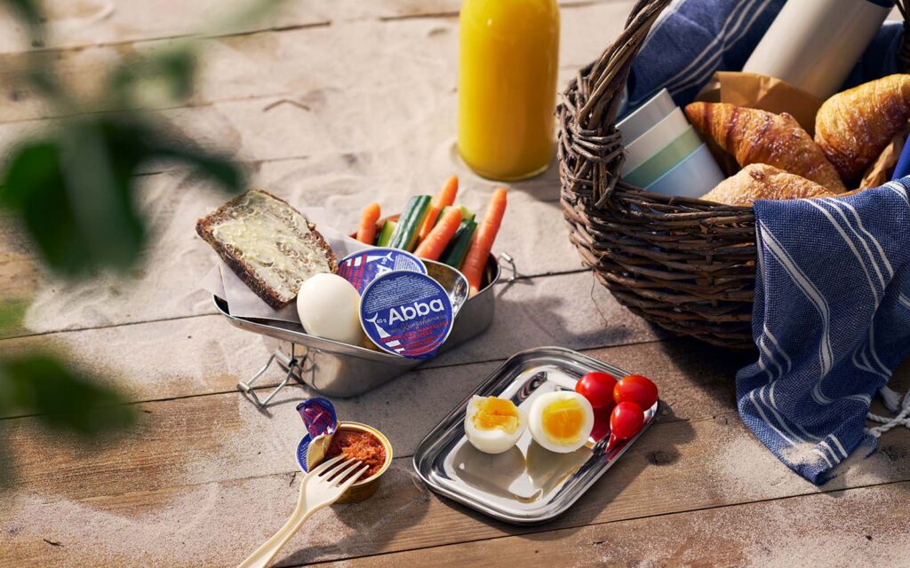 En brygga med sand och en picknickkorg. I mitten syns byttor med ägg, grönsaker, en macka och portionsförpackningar med Abba Grovhackad makrill i tomatsås.