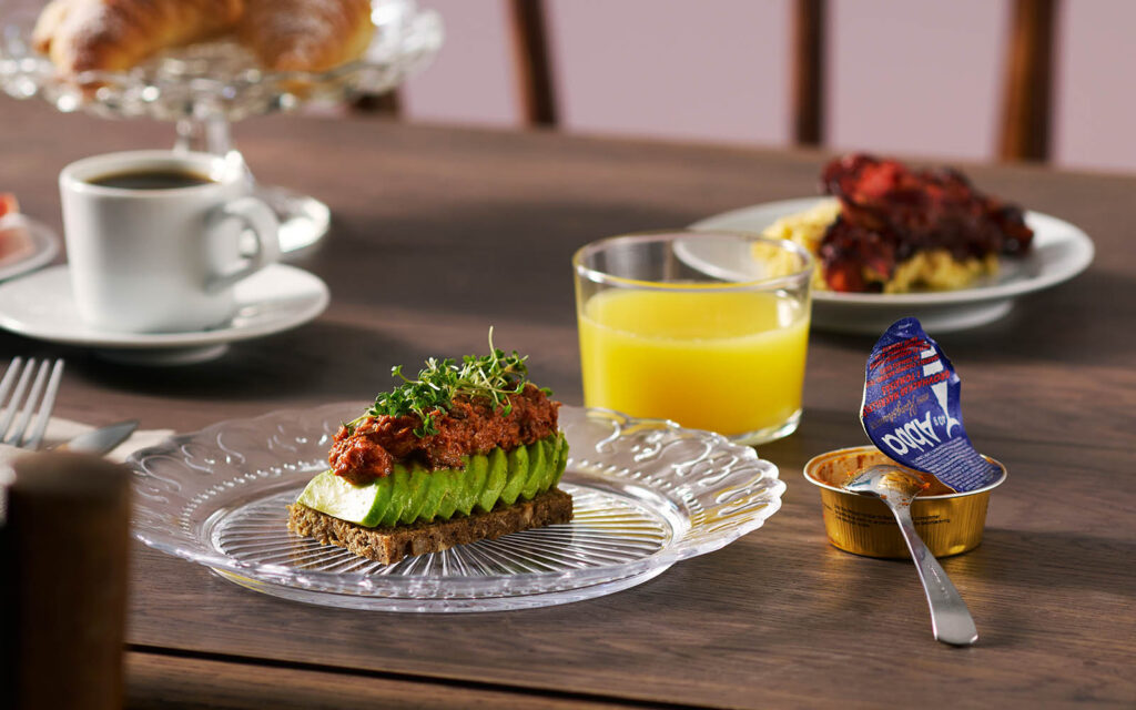 Ett fikabord med en grov macka med avokadoskivor och grovhackad makrill i tomatsås på toppen. Det står även en öppen portionsförpackning med makrill bredvid tallriken.