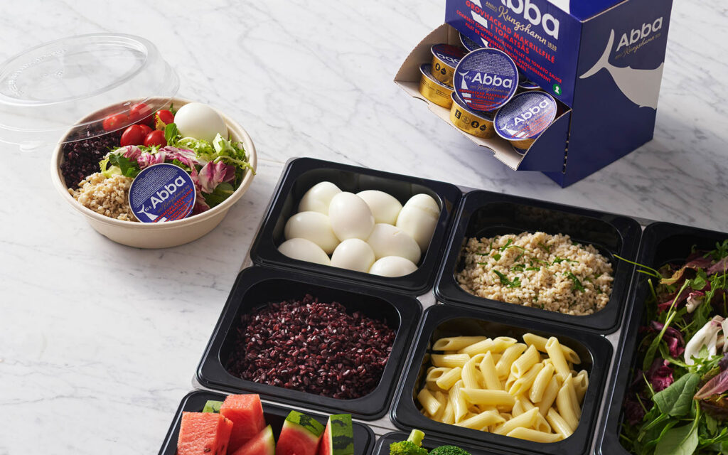 Skålar med olika ingredienser från en salladsbar, tillsammans med en färdig sallad som innehåller en portionsförpackning med Abba Grovhackad makrill. I bakgrunden syns en behållare med flera portionsförpackningar av makrillen.