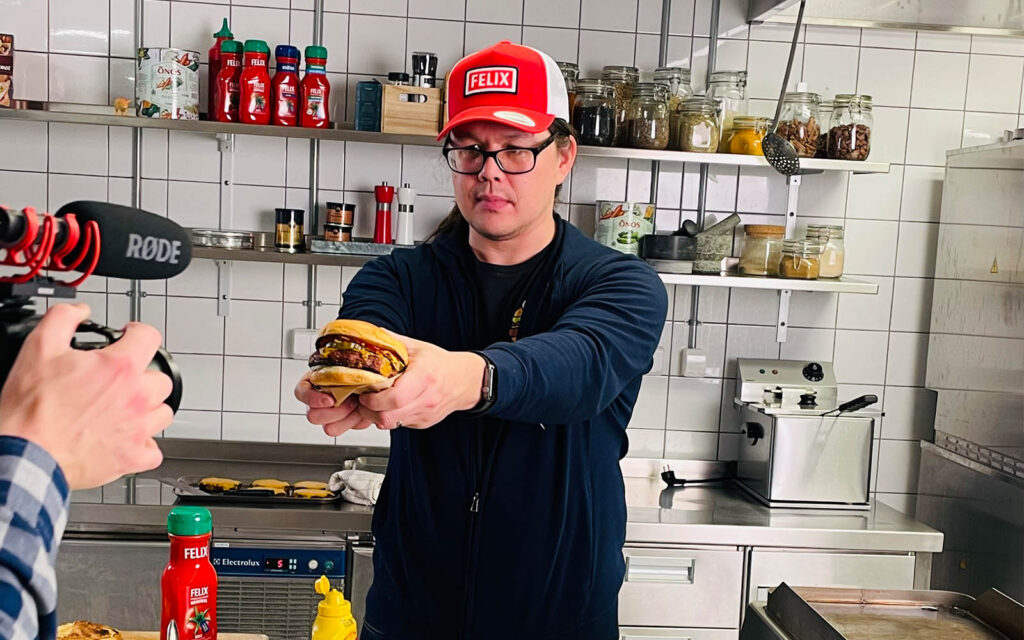 Toby Lee från Burgerdudes håller upp en burgare med två händer framför en kamera. På huvudet har han en röd keps med Felix logotyp på.