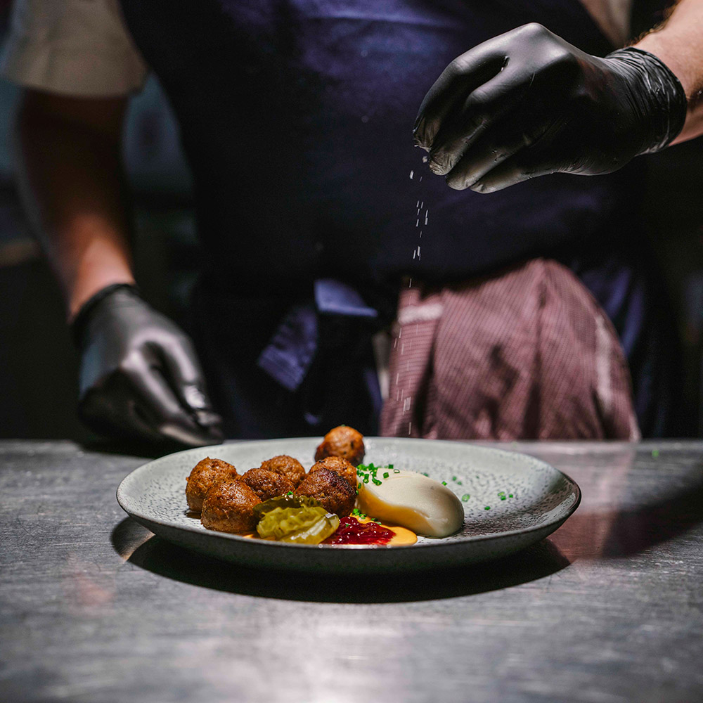 Kock med svarta arbetshandskar häller kryffa på en tallrik med köttbullar och potatismos.