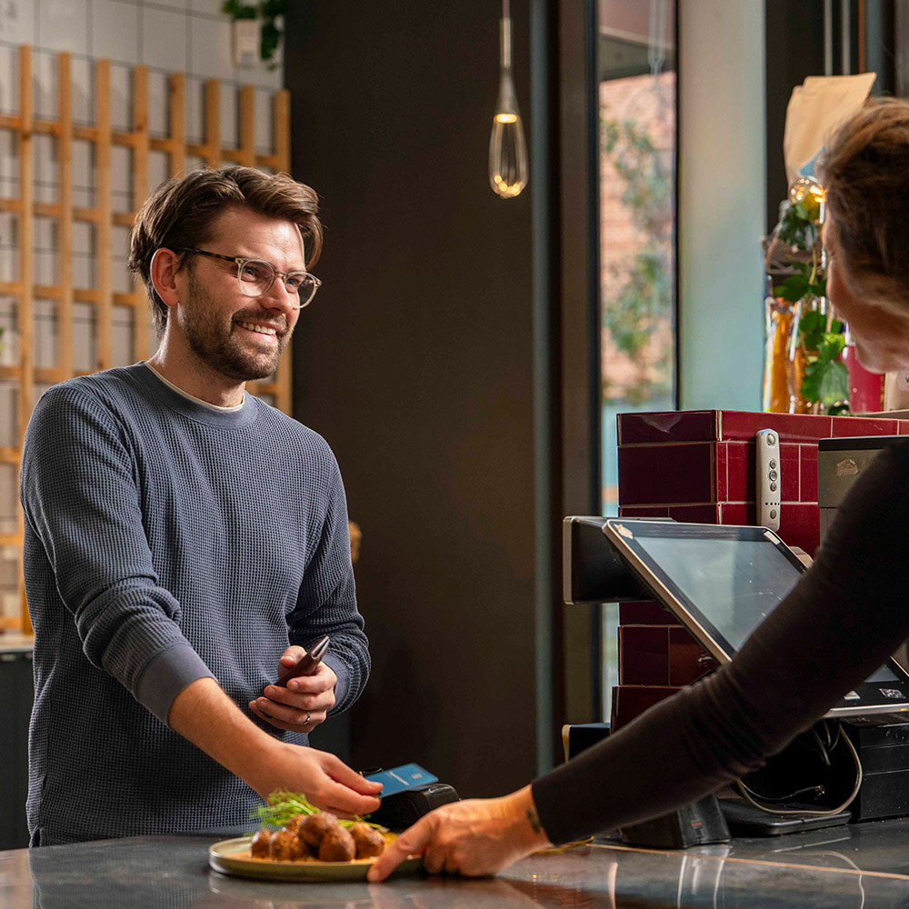 Man med blå tröja och glasögon ler vid disk och betalar med kort inne i restaurangmiljö.