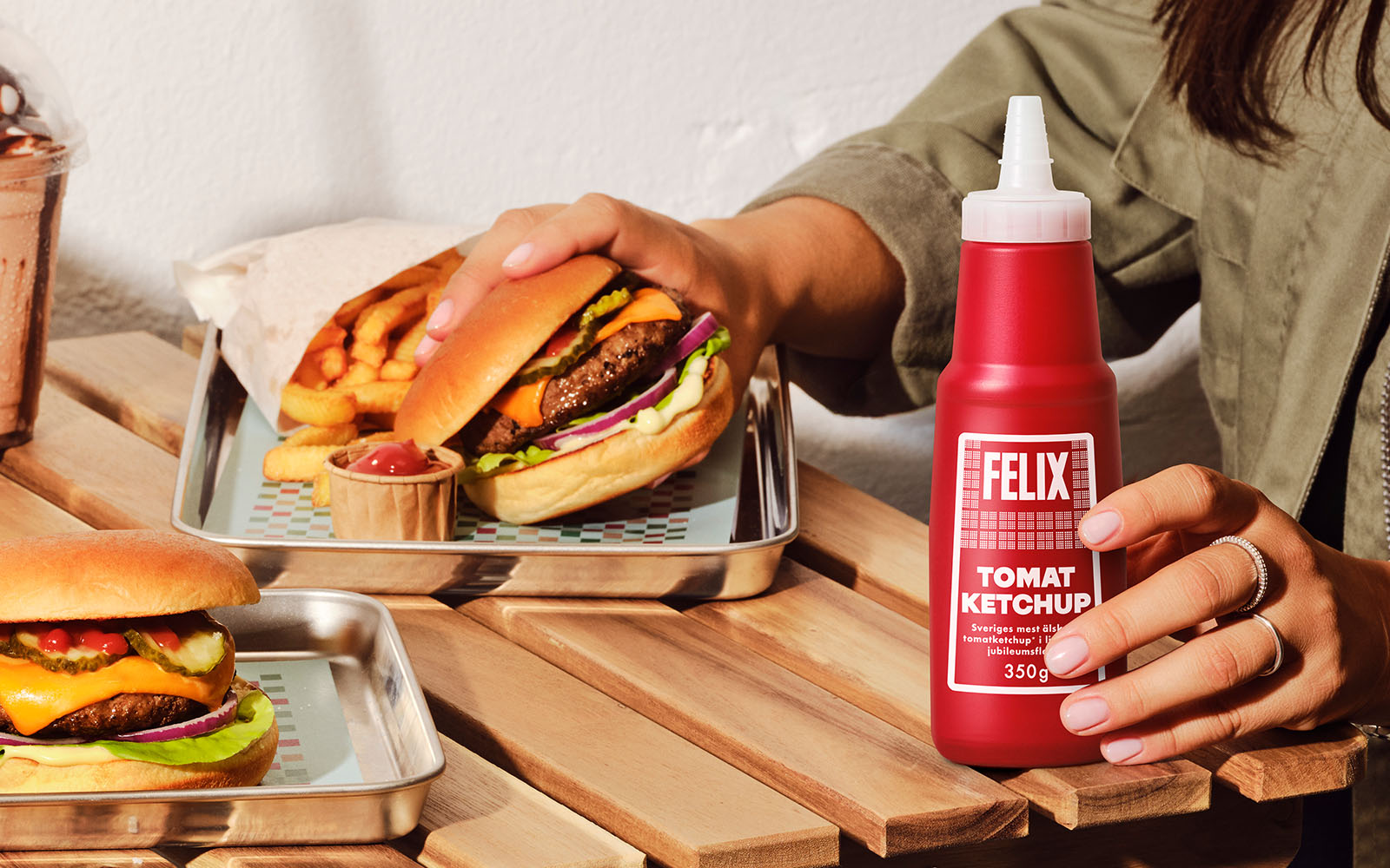 Hand som håller FELIX Tomatketchup på ett bord. I bakgrunden syns en annan hand som håller en burgare.